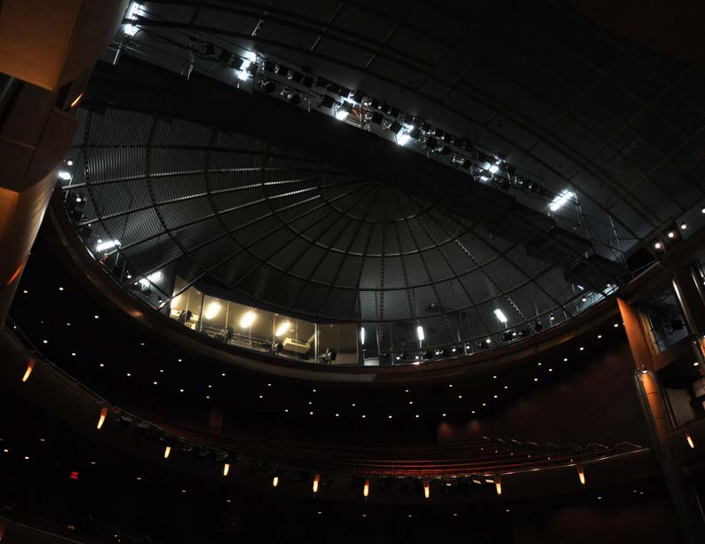 Interior view of a theater auditorium looking up at a large circular dome ceiling with stage lights and catwalks.